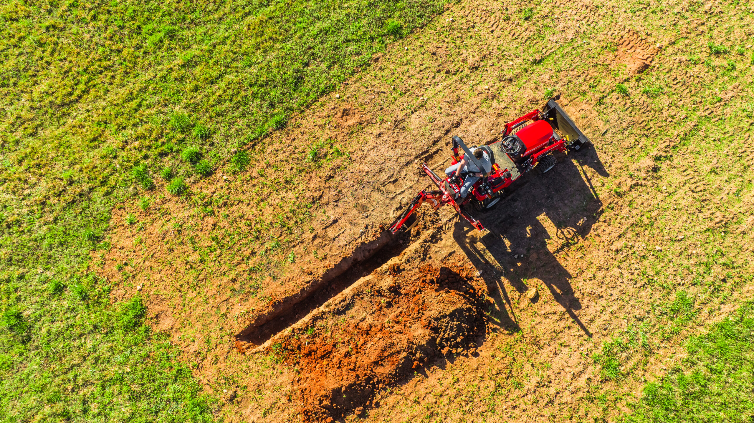Massey Ferguson 1GC Series sub-compact tractor trenching with backhoe in grassy field from aerial view