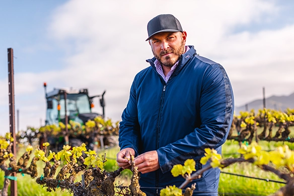 Farmer in a blue sweater standing in a vineyard with a Fendt 200 Vario tractor in the background.